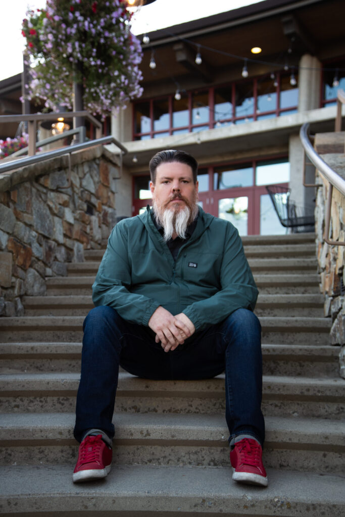 Man with a long white and brown beard sitting on outdoor stone steps wearing a green jacket, blue jeans, and red sneakers